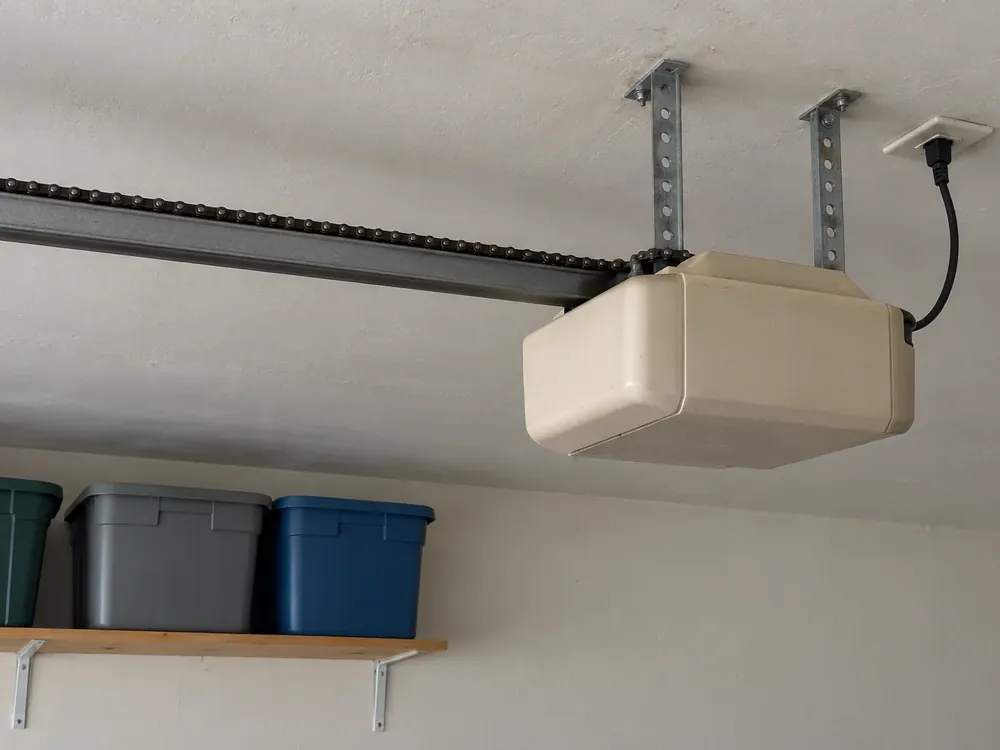 Older beige chain-drive garage door opener hung from ceiling straps, with its power cord plugged in