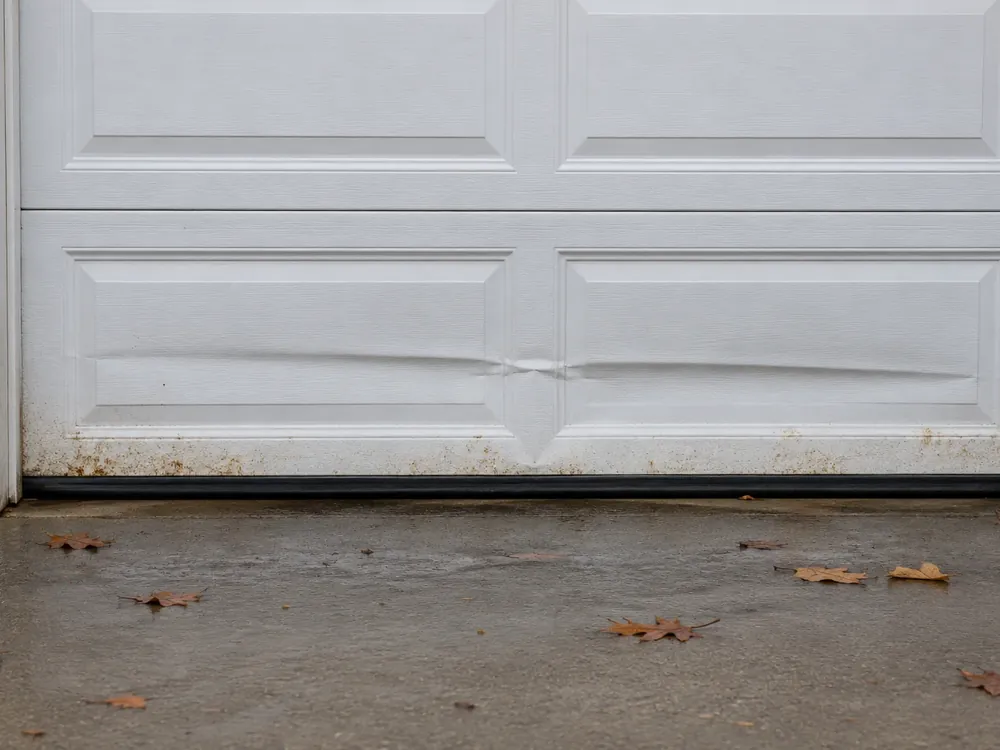 White garage door bottom section creased inward across two panels, with rust spots above the rubber seal