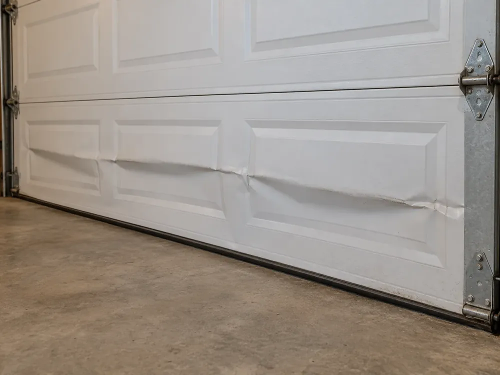 Dented lower panel of a white garage door above the black bottom seal